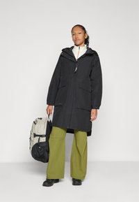Zwarte hooded parka met ritssluiting aan de voorkant, twee grote zakken en een verstelbare nek. Het model draagt olijfgroene wijde broek en heeft een grijze rugzak.