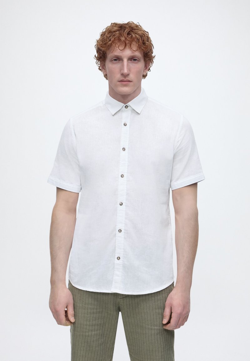 Άνδρας με κόκκινα μαλλιά, φορώντας ένα κοντομάνικο λευκό πουκάμισο με κουμπιά και πράσινο παντελόνι με ρίγες, στέκεται μπροστά σε ένα απλό ανοιχτό φόντο.