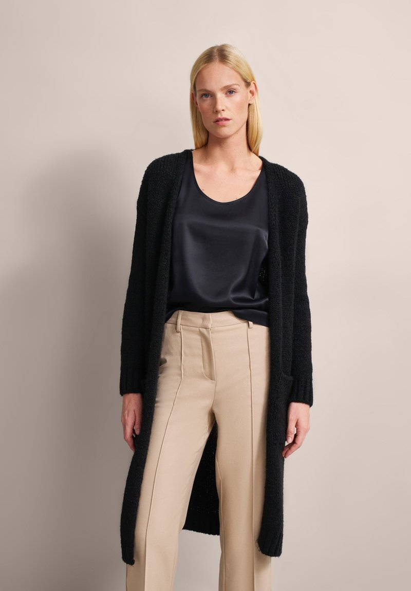 Long black cardigan with textured knit, open front, and two pockets, worn over a dark satin top and light beige tailored pants.