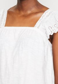 Hvid ærmeløs top med firkantet halsudskæring, der har blonder på skuldrene og et tekstureret, let stof.