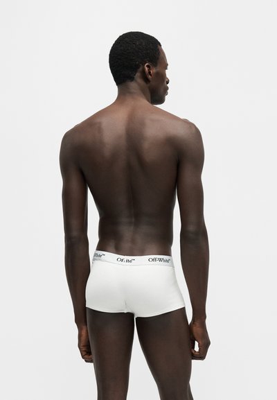 Άνδρας μοντέλο με σκουρόχρωμο δέρμα, φορώντας λευκά μπόξερ της μάρκας Off-White, στέκεται με την πλάτη προς την κάμερα σε απλό φόντο.