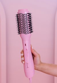 Brosse à cheveux soufflante avec un manche rose, une tête ronde, des poils noirs et un interrupteur. Finition lisse et prise texturée.