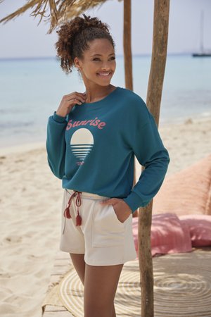 Turquoise sweatshirt met "Sunrise" grafiek en een wit cirkeldesign, gecombineerd met lichtcrème shorts met een trekkoord en zakken.