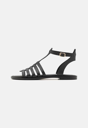Sandales en cuir noir avec design à T, présentant plusieurs sangles horizontales, fermeture à boucle sur le côté et semelle plate. Texture lisse tout au long.