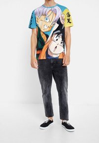 Camiseta gráfica de manga corta con personajes de anime coloridos, predominantemente azul y amarillo, combinada con pantalones gris oscuro y zapatillas negras.