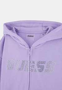 Fialový zipsový mikina vyrobená z mäkkého materiálu, s strieborným kamienkovým logom "GUESS" na prednej strane, s klasickou kapucňou a zipsovým uzáverom.