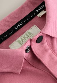 Nahaufnahme eines pinkfarbenen Hemds mit Kragen, schwarzen und weißen „Baker by Ted Baker“-Etiketten und Metall-Druckknöpfen auf der Knopfleiste.