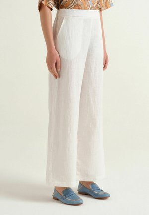 Pantaloni bianchi a gamba larga indossati con mocassini blu e una maglietta a maniche corte con fantasia, mostrati dalla metà del busto fino ai piedi su uno sfondo semplice.