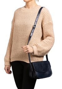 Navyblaues Leder-Crossbody-Case mit verstellbarem Riemen, runder Form, glatter Textur und kontrastierenden Nähten. Getragen mit einem strukturierten beigen Pulli.