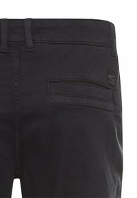 Schwarze Hose mit einer genähten Gesäßtasche und logodetail, aus einer Nahaufnahme, die die Stoffstruktur hervorhebt.