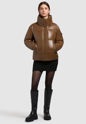 Jachetă pufosă maro, lucioasă, cu guler înalt, având panouri cusute și două buzunare frontale. Purtați-o peste o fustă neagră cu dungi și cizme negre.