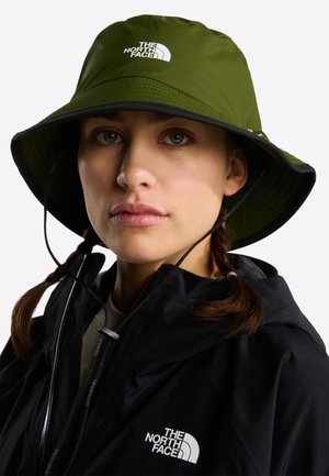 Femme portant un chapeau seau vert olive et une veste noire, toutes deux marquées du logo The North Face, regardant droit devant elle.