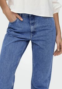 Dżinsy w średnim niebieskim odcieniu, o klasycznym prostym kroju, z lekkim wytarciem i kieszeniami z przodu. Materiał ma gładką, solidną teksturę.