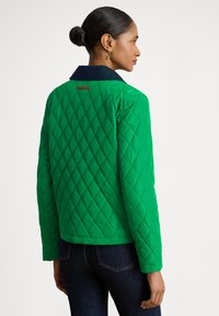 Grønn vattert jakke med diamantmønster, marineblå krage og en lærmerke bak. Har en kort passform og lange ermer.