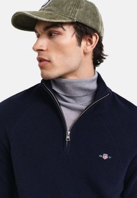 Granatowy sweter z kołnierzem na zamek o prążkowanej strukturze, nałożony na szary golf, w połączeniu z zieloną czapką z sztruksu z logo.