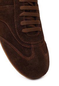 Sneaker in camoscio marrone scuro con lacci abbinati e cuciture arancioni visibili lungo la punta e i lati.