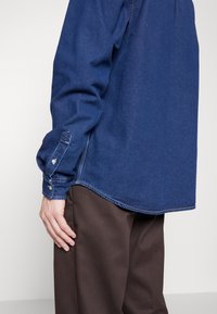 Camisa de mezclilla azul con mangas largas, puños abotonados y un dobladillo curvado. Textura suave con costuras en contraste. Llevada con pantalones marrones.