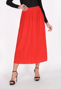 Roter plissierter Midirock mit gerafftem Bund, kombiniert mit schwarzen Riemchen-High-Heels. Glatte Textur und fließendes Design.