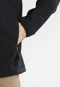 Schwarze Windjacke mit einem seitlichen Reißverschlussfach, elastischen Bündchen und einer glatten Textur. Logodetail am Ärmel.