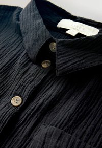 Czarna koszula z lnu o pomarszczonej fakturze, z beżowymi guzikami i spiczastym kołnierzykiem, ukazująca fakturę materiału i detale guzików.