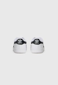 Sapatilhas brancas Adidas com um contraforte preto, apresentando uma textura de pele lisa, sola branca e logotipo em relevo no calcanhar.