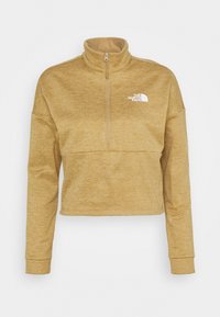 Zkrácená mikina na zip ve světle hnědé barvě, vyrobená z měkké tkaniny. Má vysoký límec a bílé logo na levé hrudi.