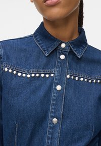Mulher a usar uma camisa de ganga azul com detalhes em tachas prateadas ao longo do peito e botões de pressão prateados na frente.