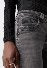 Main à la peau foncée avec une bague en or à côté d'une poche de jean grise décorée de petits clous métalliques, portant un haut noir à manches longues froncées.