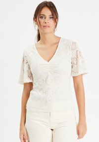 Haut en dentelle couleur crème avec un col en V, des manches courtes évasées et des motifs floraux délicats, présentant un détail de fronce à l'avant.