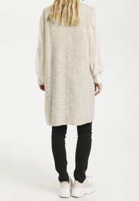 Długi, jasnoszary sweter z dzianiny o miękkiej fakturze. Noszony na przezroczystą, długą bluzkę, w komplecie z czarnymi spodniami i białymi trampkami.