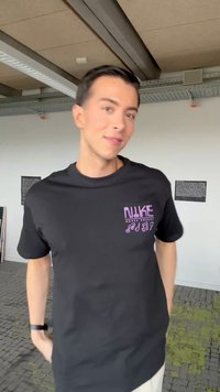 Fiatal férfi fekete "NIKE Music Energy" pólóban áll beltéren, kezeit zsebekben tartja, enyhén mosolyog, a látható mennyezeti gerendák alatt.