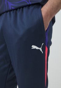 Jūras spēku sportiskās bikses, kas izgatavotas no gluda materiāla, ar baltu Puma logotipu sānos un spilgti rozā akcentu uz augšstilba.