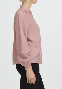Hellrosa Bluse mit langen Ärmeln aus strukturiertem Stoff, mit Stehkragen und gerafften Manschetten, getragen zu schwarzen Hosen.