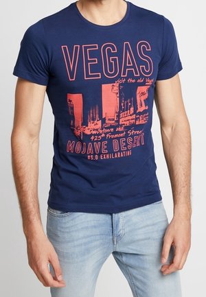 Homme portant un t-shirt bleu marine avec un texte graphique rouge et des images faisant référence à Vegas, au désert de Mojave et à Fremont Street.