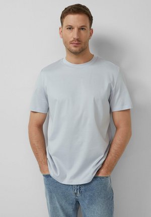 Mężczyzna stoi oparty o szarą ścianę, ubrany w jasnoniebieską koszulkę z krótkim rękawem i niebieskie dżinsy, z rękami w kieszeniach.