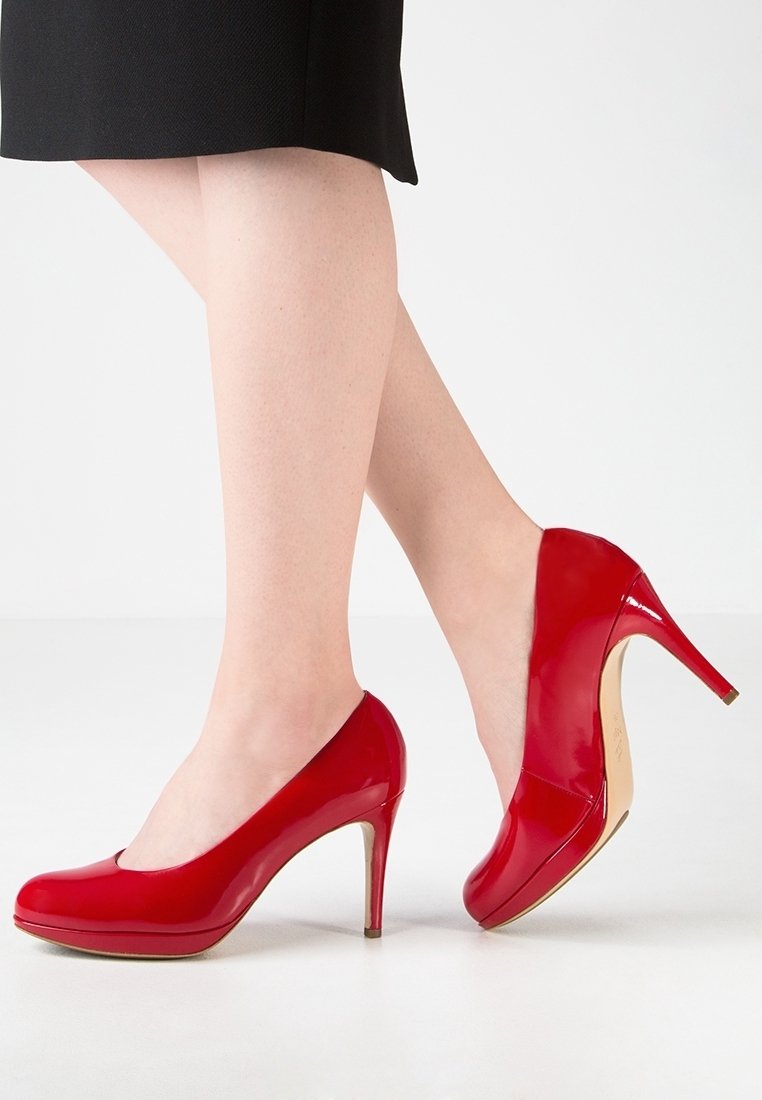 Femme portant des chaussures à talons hauts rouges brillantes avec une jupe noire, debout devant un fond blanc uni.