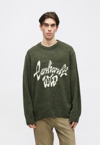 Pulover tricotat de culoare verde khaki, cu guler rotund și croială lejeră, având un text grafic alb pe față. Textură moale și mâneci lungi.
