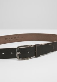 Brun læderbælte med en sølvmetalspænde, prægede logoer og flere justeringshuller for størrelsesfleksibilitet.