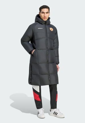 Nuori mies pukeutuneena mustaan Adidasin toppatakkiin, jossa on huppu ja Manchester Unitedin logo, musta-puna-valkoiset housut ja valkoiset Adidas-tennarit.