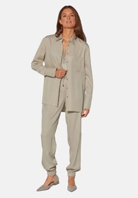 Helles Beige, Langarmhemd mit Tasche, kombiniert mit passenden, schmal zulaufenden Hosen. Weicher Stoff; texturierte Details am Oberteil, abgerundet mit grauen Schuhen.