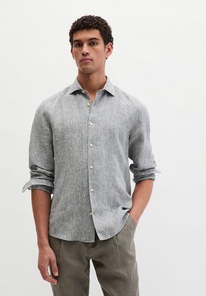 Jonge man met krullend haar, gekleed in een lichtgrijs linnen overhemd met opgerolde mouwen en olijfgroene broek, staand tegen een effen achtergrond.