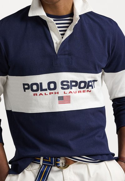 Jūras zilas regbija krekls ar baltu horizontālu svītru, kurā ir uzraksti "POLO SPORT" un "RALPH LAUREN", kā arī amerikāņu karoga ikona. Krekls ar apkakli.