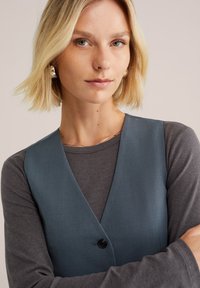 Vesta albastră peste un tricou gri cu mânecă lungă, având un decolteu adânc în V și o închidere cu nasturi, cu o textură fină și un ușor luciu.