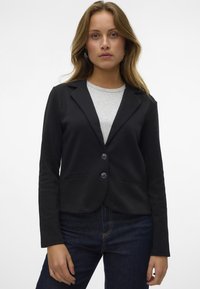 Černý látkový blazer se dvěma knoflíky, klopami a bočními kapsami, spárovaný se světle šedým topem a tmavými džínami. Hladká textura.