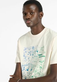 T-shirt en coton crème avec un imprimé graphique bleu représentant des figures actives et des scènes liées au sport, col rond et manches courtes.