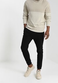 Helles beigefarbenes Sweatshirt mit strukturiertem Oberteil, kombiniert mit schwarzen, schmal zulaufenden Hosen und beigen Sneakers mit weißen Akzenten.