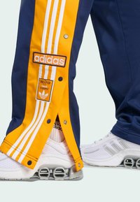 Blue and yellow Adidas track pants with snap buttons, featuring three white stripes and a brand logo patch, paired with white sneakers.