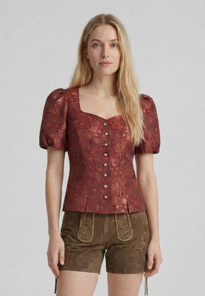 Frau mit langen blonden Haaren, die eine rote gemusterte Bluse mit Puffärmeln und braune bestickte Lederhosen trägt, steht vor einem schlichten Hintergrund.