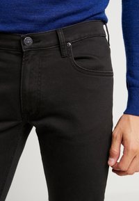 Närbild av en person som bär svarta jeans med en metallknapp från Lee och en blå långärmad topp, med handen vilande vid sidan.