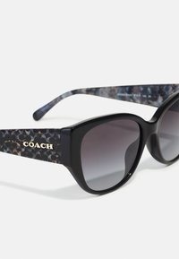 Schwarze Sonnenbrille mit dickem Rahmen, Verlaufslinsen und strukturierten Acetat-Bügeln mit einem dunklen Muster und goldenem "COACH"-Logo.